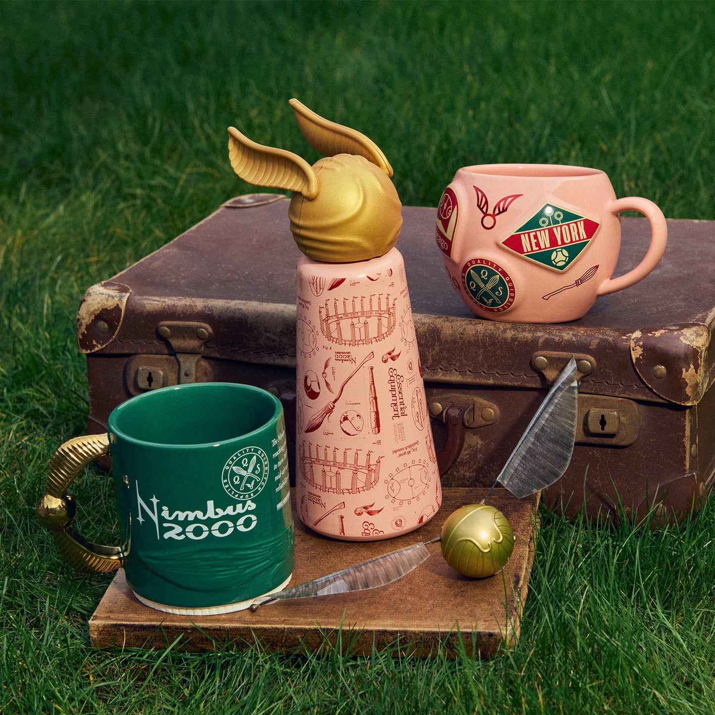 Decorative items including a pink Quidditch mug, a golden snitch water bottle, and a green Nimbus 2000 mug on a grassy background.