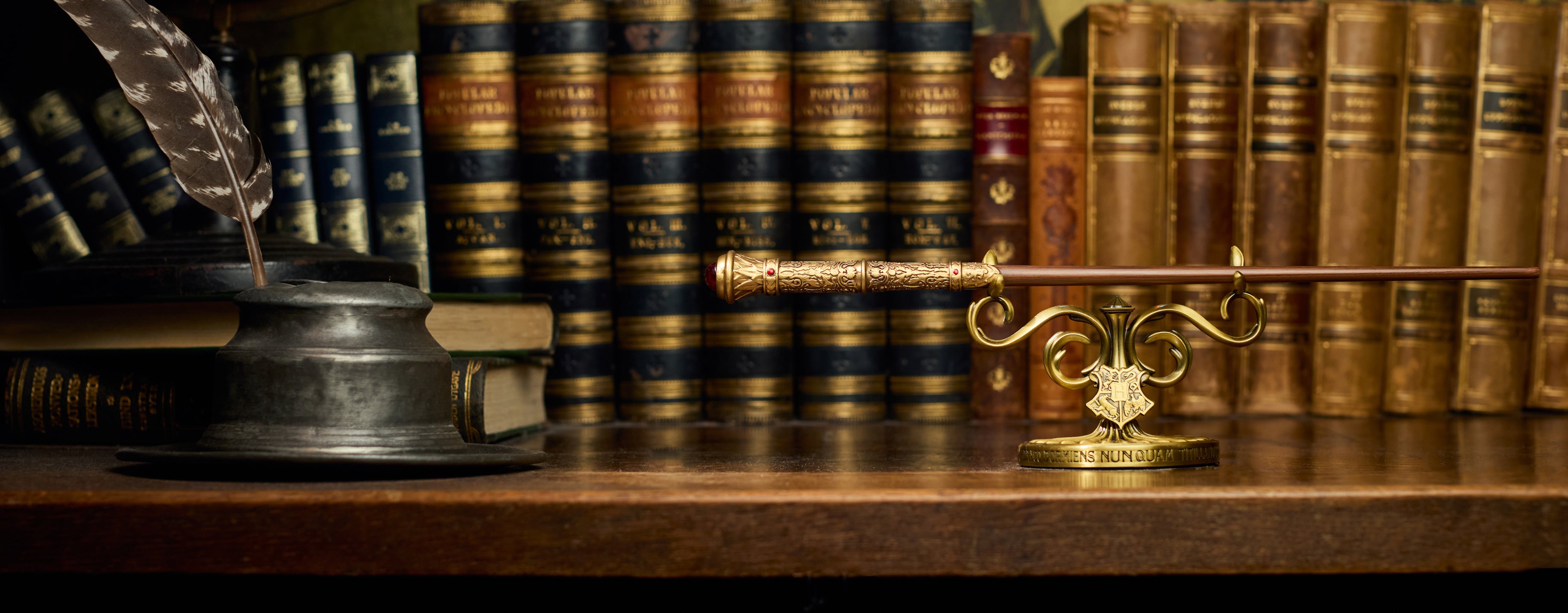 A gold ornate wand stand rests on a wooden desk in front of a row of antique leather-bound books, alongside a dark inkwell and quill.