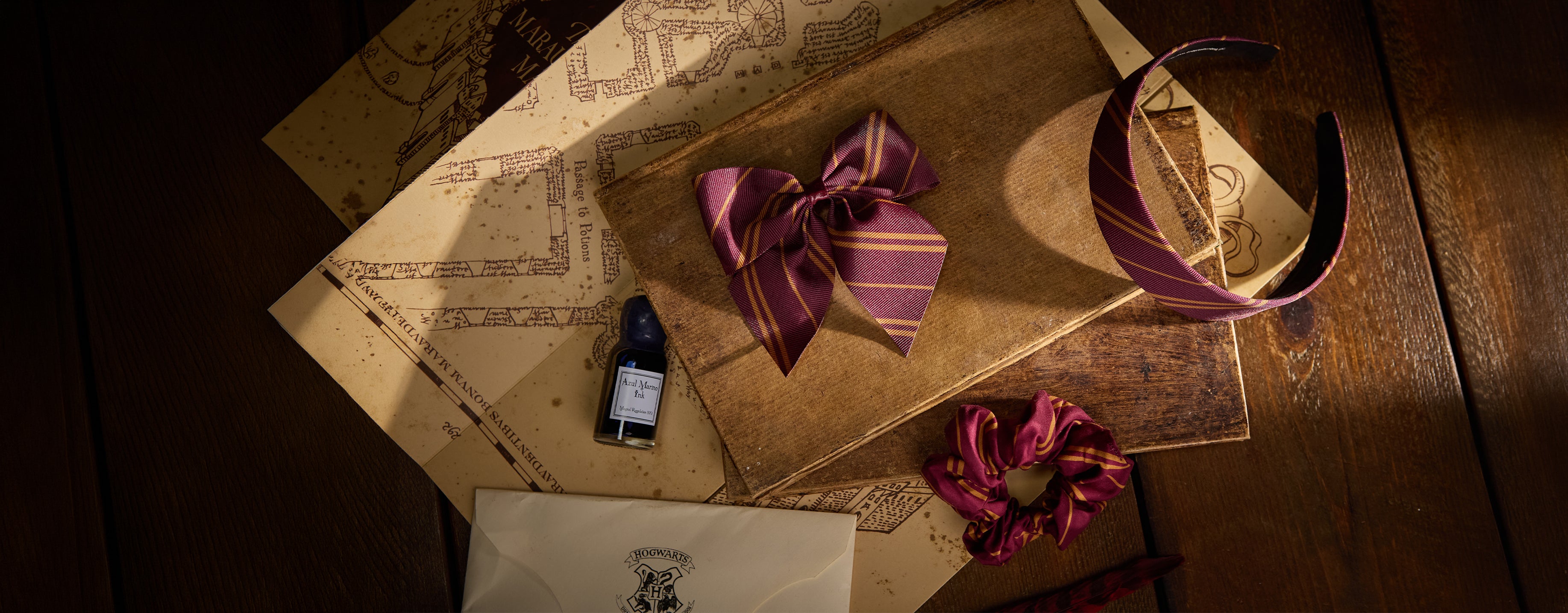 A styled flat-lay of Harry Potter themed accessories on a wooden table, including a burgundy and gold striped bow, matching scrunchie, headband, and small bottle, arranged with the Marauder's Map and acceptance letter.