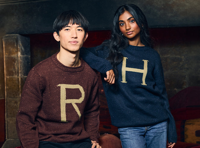 Two models wearing knit sweaters with large letter graphics, posed in front of a dark fireplace. The model on the left wears a brown sweater with a gold 'R', and the model on the right wears a navy blue sweater with a gold 'H'.