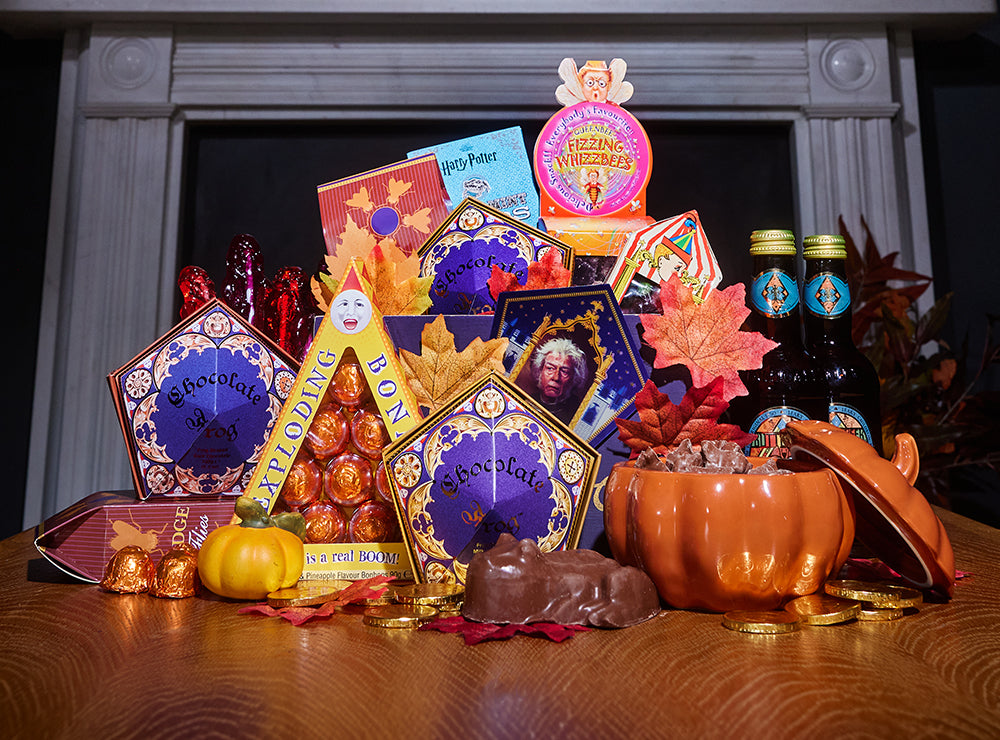 A styled assortment of Halloween-themed chocolates, candies, and treats arranged on a wooden table in front of a fireplace. The display includes branded boxes and packages such as 'Chocolate Frog', and 'Fizzing Whizzbees', alongside decorative fall leaves, a small pumpkin, chocolate coins, and an orange pumpkin-shaped container.