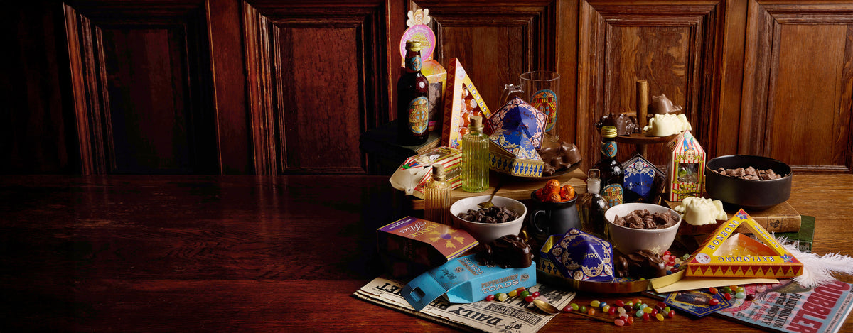 A styled tabletop arrangement of assorted candy, snacks, small bottles, and packaged treats set against a dark wood paneled background.