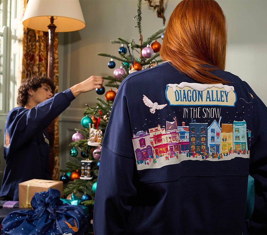 A person with red hair stands with their back to the camera wearing a navy blue sweatshirt featuring a colorful illustrated holiday town scene and the text 'DIAGON ALLEY IN THE SNOW.' Another person is visible in the background decorating a Christmas tree in a cozy indoor setting.