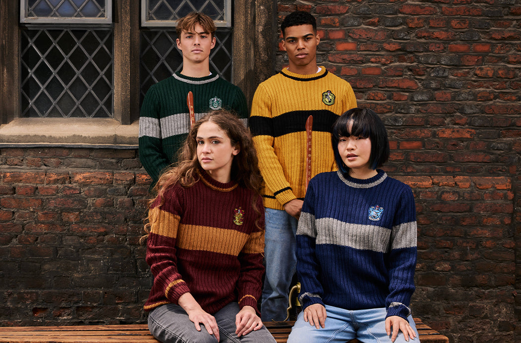 Four young adults wearing Hogwarts-themed striped knit sweaters pose together in front of a rustic brick wall. The sweaters feature the Gryffindor, Slytherin, Hufflepuff, and Ravenclaw crests and are styled with wooden wand props.