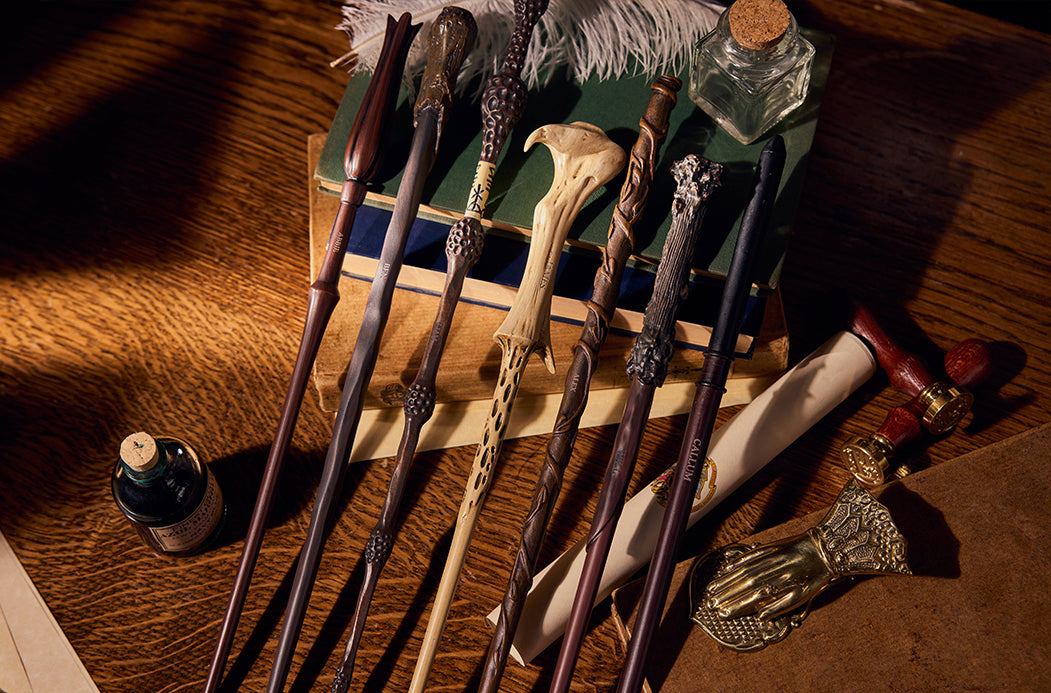 A set of seven intricately carved and shaped wizard's wands arranged on a wooden desk alongside themed props including old books, a small ink bottle, a scroll, and a miniature hand-shaped object.