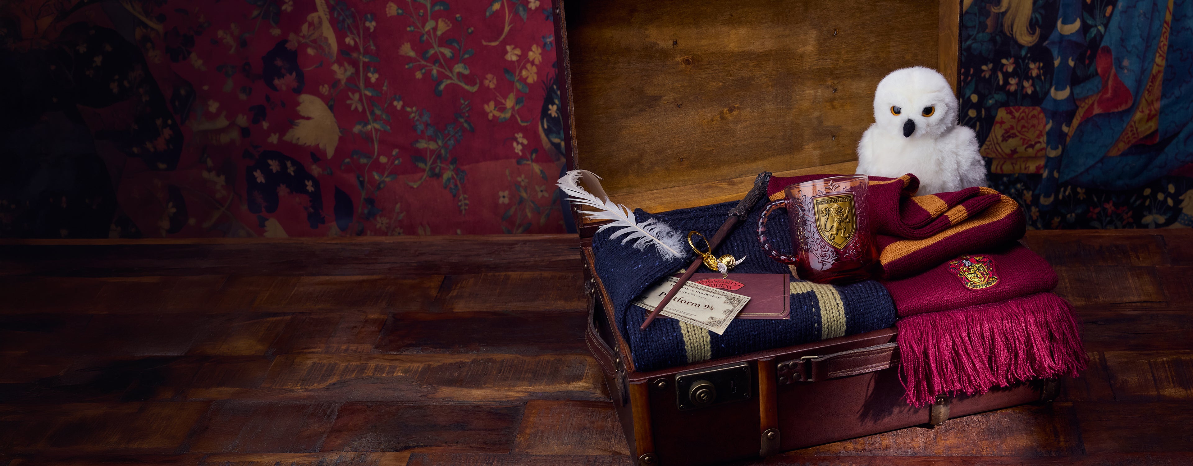 Harry Potter Gift Trunk filled with items including a mug, soft toy, scarf, wand, keyring, feather, Platform 9 3/4 ticket, on a wooden floor