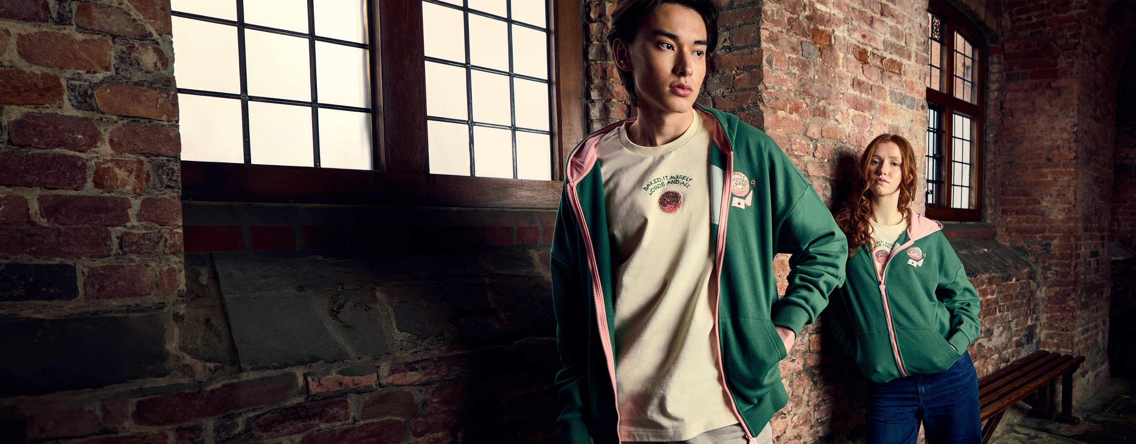Two people wearing green hoodies in a brick-walled corridor with large windows.