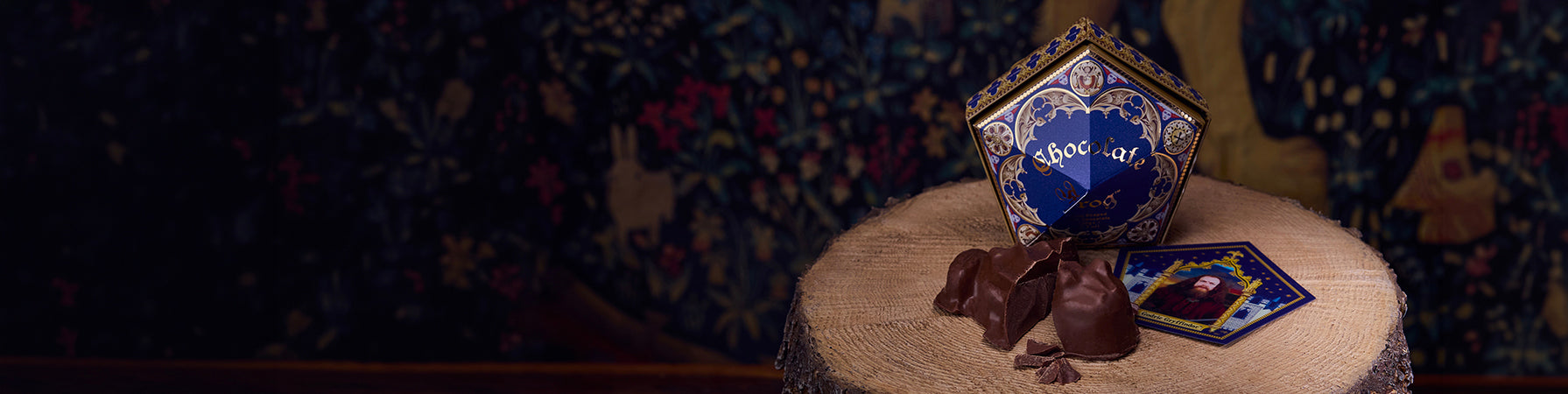 A decorative blue and gold Honeydukes chocolate frog box sits on a round wooden stump next to broken dark chocolate pieces and a small matching card, against a dark patterned tapestry background.