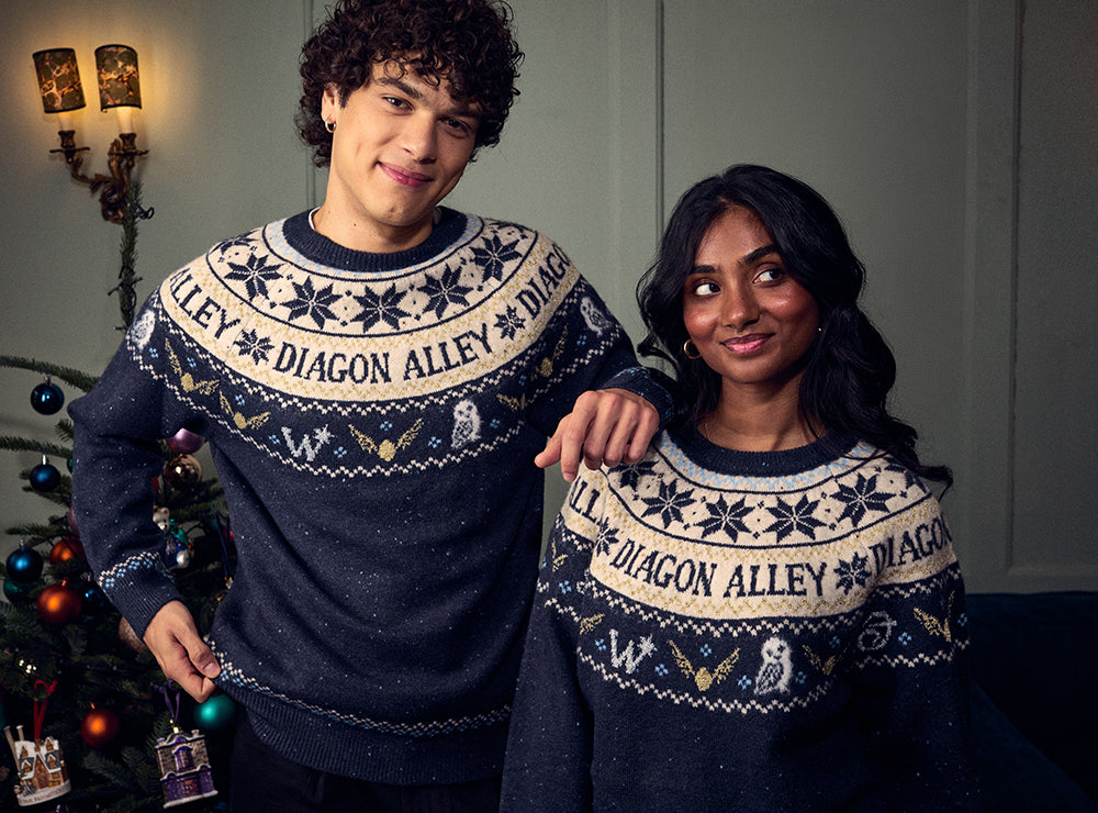 Two people wearing matching navy and cream holiday sweaters with 'DIAGON ALLEY' and owl-themed graphics, posed in front of a decorated Christmas tree and wall sconces.