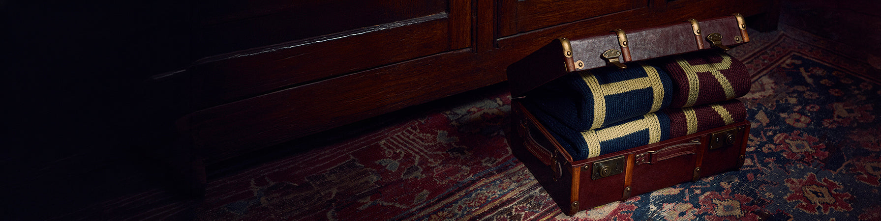 A vintage brown leather suitcase with brass hardware sits open on a patterned red and blue rug next to a dark wooden cabinet in a dimly lit room.