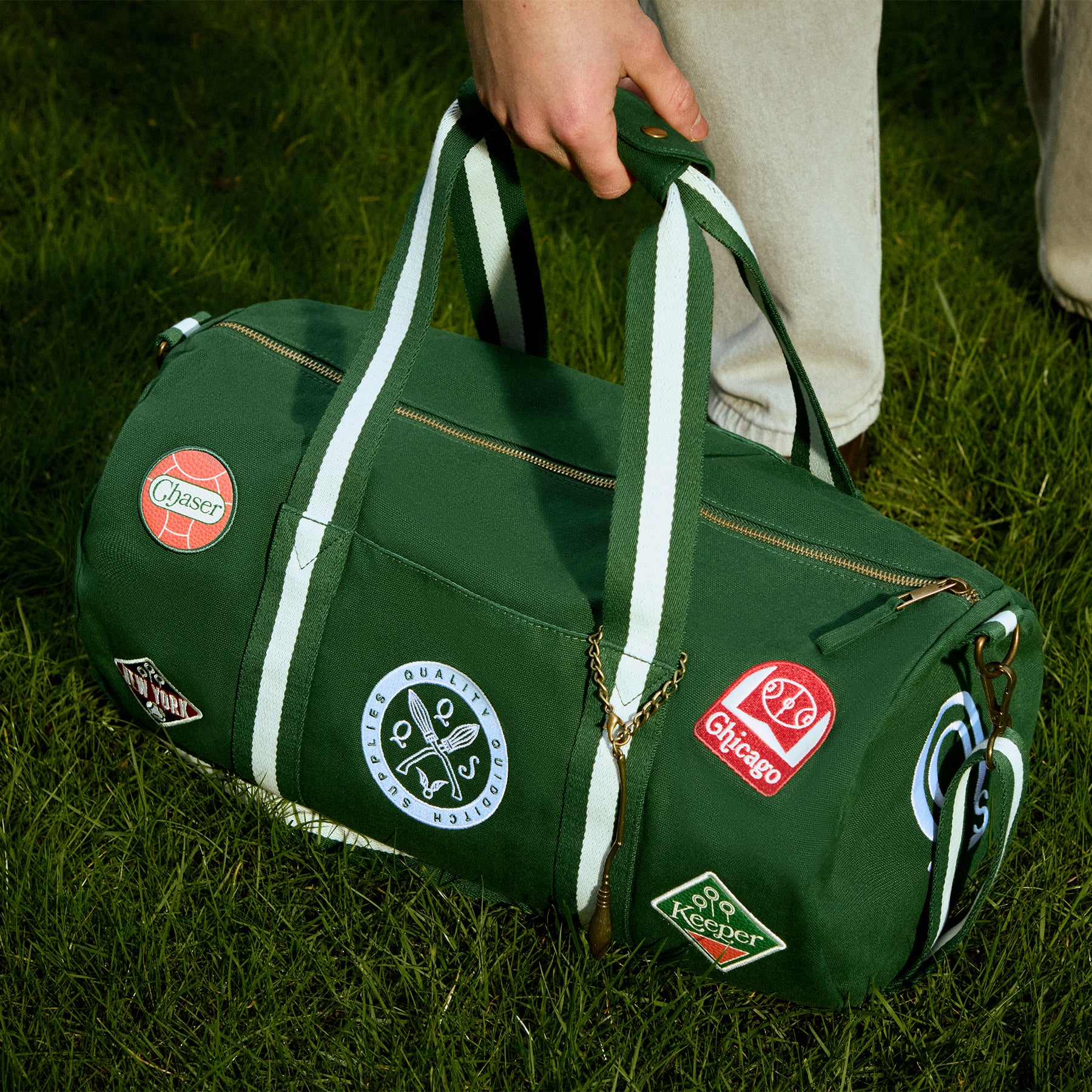 Green duffel bag with various Quidditch patches held by a person on grass