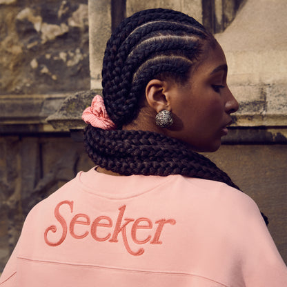 Person wearing a pink sweatshirt with 'Seeker' printed on the back and a pink Quidditch scrunchie, standing against a stone wall.