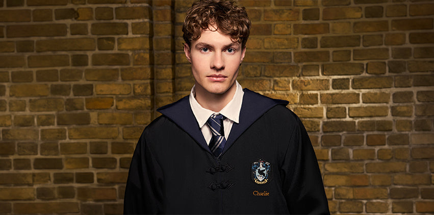 A young man with curly brown hair wearing a Hogwarts robe with a white shirt and striped tie, standing against a warm brown brick wall. The robe features a Ravenclaw crest on the left chest and the name 'Charlie' embroidered below it.