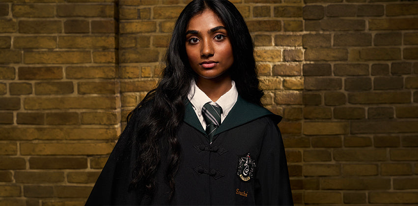 A young woman with long dark hair stands against a warm brick wall wearing a dark Hogwarts-style robe with a green collar, white shirt, and striped tie. A crest patch is visible on the left chest, and her name appears embroidered on the right side.